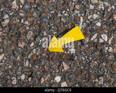 une petite flèche métallique jaune avec des traces d'utilisation se trouve sur une surface de route accidentée et pointe vers le bas à gauche Banque D'Images