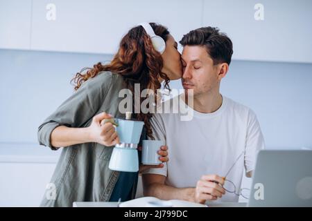 Une femme aux cheveux longs et bouclés sourit dans un casque et verse du café dans une tasse pour un mari stressé travaillant sur un ordinateur portable et résolvant les problèmes de travail Banque D'Images