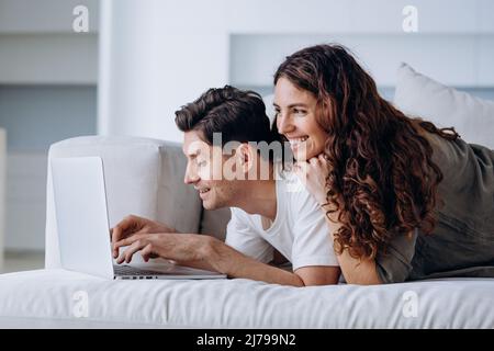 Couple amoureux se trouve sur le canapé derrière un ordinateur portable dans le salon près de la fenêtre et fait des achats en ligne ou prévoit un voyage Banque D'Images