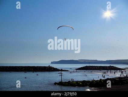 Un parapente motorisé au-dessus des rochers de Chit près de Jacobs Ladder Beach Sidmouth, East Devon pris dans le soleil rétro-éclairé. Les jeunes et les personnes âgées en dessous apprécient Banque D'Images