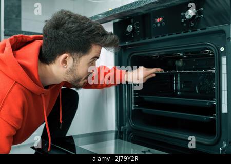 Four intégré, le jeune homme ouvre la porte du four et prépare le repas, le beau jeune homme prépare sa propre nourriture Banque D'Images