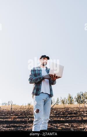 Jeune agriculteur latin homme teleworking sur son ordinateur portable au milieu de ses terres agricoles . Durabilité agricole Banque D'Images