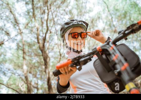 fille asiatique voilée tenant des lunettes de soleil souriant tout en faisant du vélo dans le parc Banque D'Images