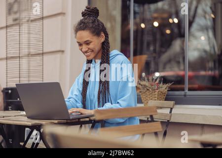 Bonne jeune jolie femme élégante assise sur la terrasse du café et textant sur un ordinateur portable. Indépendant. Banque D'Images