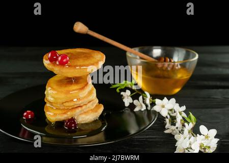 Crêpes au miel sur fond noir avec une branche de cerisiers en fleurs. La main tient un bâton de bois versant un jet de miel. La lévitation alimentaire. Banque D'Images