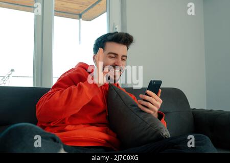 FaceTime, le jeune homme, discute vidéo avec son ami et lui fait signe, un jeune adulte assis sur un canapé dans le salon ayant une détente et un plaisir Banque D'Images