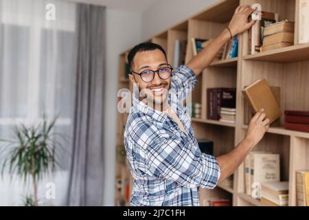 Bibliothèque personnelle. Un homme arabe heureux prenant livre de la bibliothèque, la recherche et le choix manuel, souriant à l'appareil photo, l'espace de copie Banque D'Images