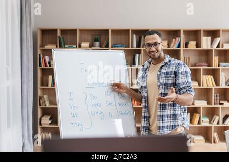 Formation en ligne. Portrait d'un homme arabe joyeux enseignant pointant sur un tableau blanc et expliquant les règles de grammaire anglaise Banque D'Images