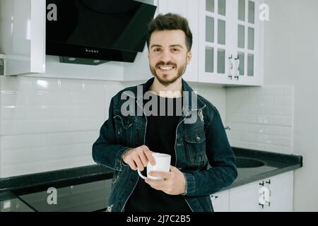 Pose, jeune homme souriant et tenant une tasse dans la cuisine, adulte homme portant une veste en denim posant pour un appareil photo Banque D'Images