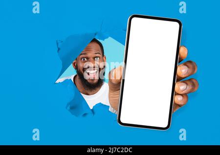 Un gars enthousiaste montrant un écran vide blanc de smartphone à travers du papier déchiré Banque D'Images