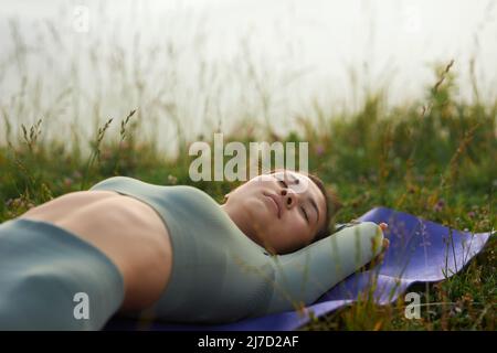 Vue latérale de la femme en vert dormir à l'extérieur après avoir fait des exercices de yoga. Récolte de femme se détendant paisiblement après l'exercice de yoga parmi l'herbe tenant les mains derrière la tête. Concept de calme. Banque D'Images
