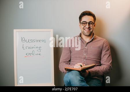 Professeur avec des lunettes tenant un livre dans sa main assis à la maison sur une chaise à côté d'un tableau blanc qui lit l'éducation, l'apprentissage, l'étude. Mur blanc Banque D'Images