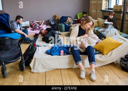 Jeune mère attentive en tenue décontractée bercer son bébé enveloppé dans un tissu écossais chaud tout en étant assise dans un endroit où dormir dans un abri pour les réfugiés Banque D'Images