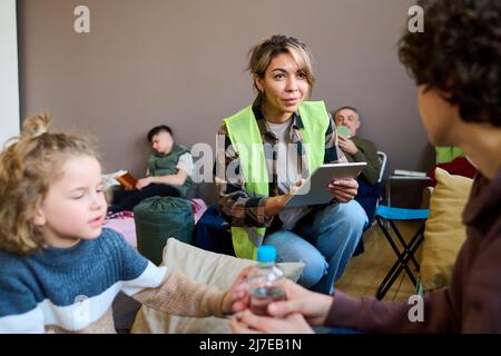 Jeune femme volontaire avec tablette assis sur les squats devant la femme avec son fils et en entrant leurs noms et autres données personnelles Banque D'Images