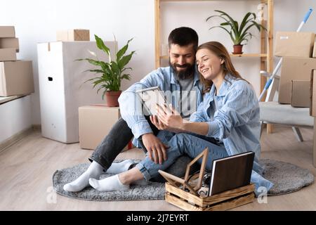 un jeune couple heureux s'assoit sur le sol dans des vêtements décontractés qui embrasent la photo encadrée. Une pile de boîtes en carton et de plantes de maison près d'un mur blanc à l'intérieur Banque D'Images