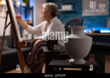 Gros plan d'un vase blanc sur une table en bois utilisé pour l'inspiration par un artiste âgé qui esquisse sur un chevalet dans un studio d'art créatif. Accent sélectif sur la durée de vie fixe utilisée comme sujet pour le dessin en atelier. Banque D'Images