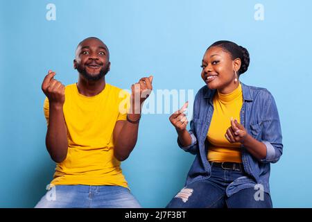 Couple faisant le geste d'argent et exigeant des récompenses d'argent pendant qu'ils sont assis dans le studio. Homme et femme se frottant les doigts pour demander le paiement, se sentant optimiste. Personnes utilisant le langage corporel pour recevoir des revenus. Banque D'Images