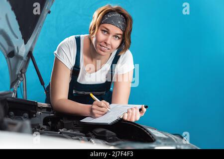 Le portrait d'une jeune femme de mécanicien caucasien en combinaison signe des documents se penchant à la voiture. Le concept de l'égalité de la femme et travaille à la réparation automatique s. Banque D'Images