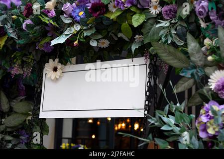 Scène de maquette d'affiche de tableau de mariage vierge. Tableau d'accueil blanc vide avec fleurs. Guirlande fleurie sur arc, décoration d'anniversaire. Banque D'Images