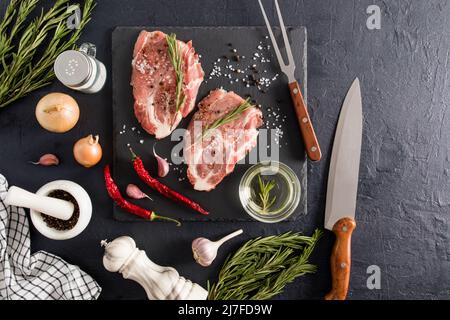 deux steaks de porc crus sur une plaque d'épices en pierre entourée d'ingrédients pour la cuisson. couteau, fourchette pour faciliter la coupe. délicieux repas Banque D'Images