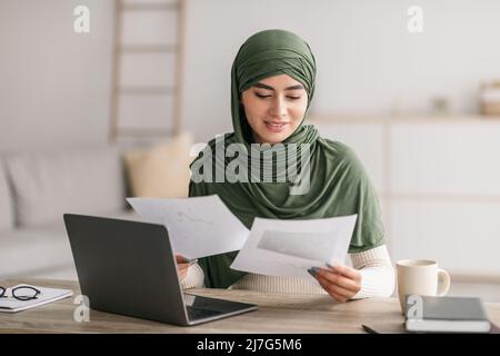 Une jeune femme arabe heureuse dans le hijab utilisant un ordinateur portable, regardant à travers les documents d'affaires pendant la réunion de travail en ligne à la maison Banque D'Images