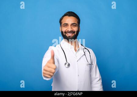Joyeux médecin indien avec un stéthoscope dans une robe médicale posant isolé sur fond bleu, montrant un pouce vers le haut. Un professionnel de la santé souriant et accueillant qui regarde la caméra Banque D'Images