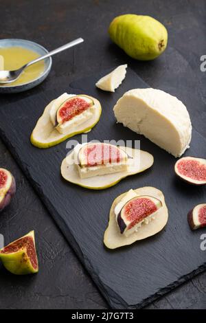 Apéritif d'été avec poire, fromage cottage, figues et miel sur panneau d'ardoise sur fond de béton noir. Vue latérale, gros plan. Banque D'Images