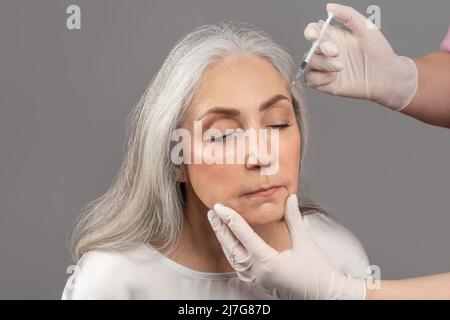 Charmante femme âgée recevant une injection de beauté, recevant de l'acide hyaluronique ou du collagène sur fond gris studio Banque D'Images