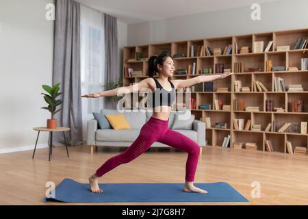 Concept de yoga domestique. Jeune femme asiatique mince faisant une fente, debout dans l'asana guerrier pendant l'entraînement à la maison, pleine longueur Banque D'Images