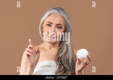 Belle femme sénior appliquant de la crème hydratante sous ses yeux sur fond de studio brun Banque D'Images