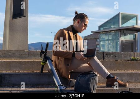 Homme d'affaires assis sur les escaliers de la ville à l'aide de sa tablette électronique avec le scooter électrique penché derrière lui à l'heure du coucher du soleil Banque D'Images