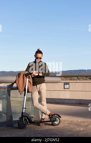Vue verticale d'un jeune homme moderne qui se moque d'un scooter électrique dans le parc et se divertit avec la nouvelle montre intelligente Banque D'Images