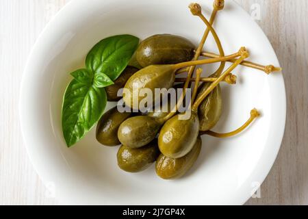 Fruits de câpres marinés et feuilles de basilic vert dans un bol blanc. Baies de câpres marinées. Caberberry en conserve comme garniture et condiment. Cuisine méditerranéenne. Banque D'Images