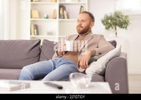 Jeune homme assis sur un canapé à la maison avec une tasse de thé et de réflexion Banque D'Images