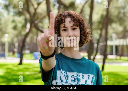 Jeune femme à tête rouge portant un t-shirt vert montrant le numéro un avec le doigt avec un peu de sourire, à l'extérieur. Attention sélective sur son visage. Banque D'Images