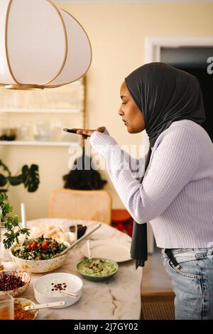 Une jeune femme en hijab photographiant de la nourriture à travers un smartphone à la table de salle à manger Banque D'Images