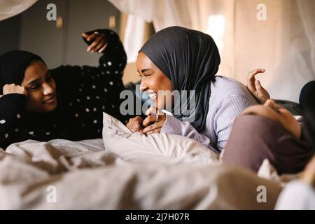 Jeune femme gaie portant le hijab parlant avec des amis au lit à la maison Banque D'Images