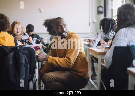 Vue latérale d'un enseignant parlant avec un élève tout en se croquant au milieu des bureaux dans la salle de classe Banque D'Images