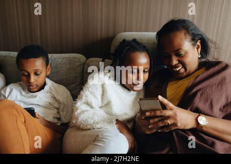 Femme souriante utilisant un smartphone tout en étant assise avec sa fille et son fils sur le canapé à la maison Banque D'Images