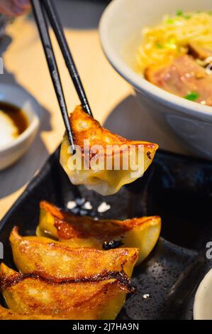 Les baguettes ramassent des gyoza frits (boulettes) de style japonais d'une assiette noire. Banque D'Images