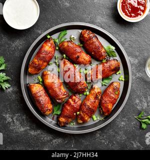 Ailes de poulet grillées enveloppées de bacon sur une assiette sur fond de pierre sombre. Vue de dessus, plan d'appartement Banque D'Images