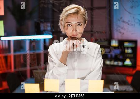 Une femme d'affaires intelligente et créative utilise des notes adhésives dans un mur de verre pour élaborer un plan d'affaires stratégique. Jeune femme caucasienne travaillant au bureau dans la soirée, debout devant un panneau de verre. Banque D'Images