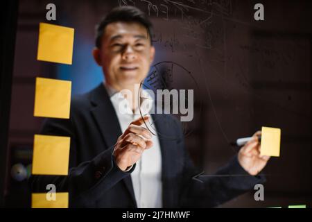 Vue rapprochée floue d'un beau homme d'affaires asiatique souriant, debout derrière un mur de verre de bureau, en utilisant des notes de poste tout en planifiant des tâches de projet dans un bureau moderne la nuit. Concentrez-vous Banque D'Images