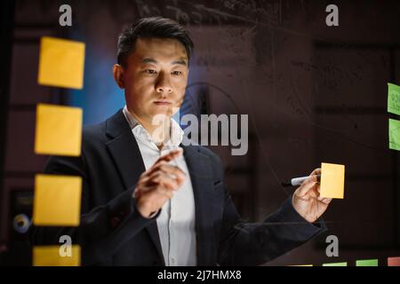 Regardez à travers le panneau de verre de l'homme d'affaires asiatique concentré, en pensant aux façons de résoudre les problèmes, en attachant des feuilles de notes de couleur sur le mur de verre et en traçant les tableaux Banque D'Images