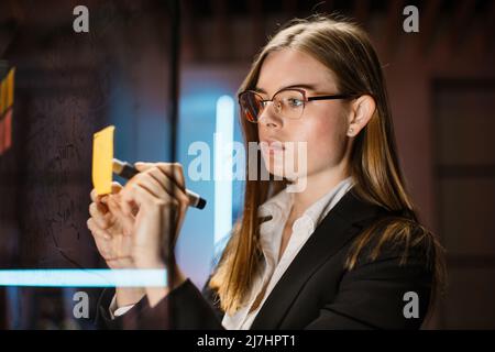 Une jeune femme d'affaires écrit des tâches ou des idées créatives sur un post-it collant sur un tableau transparent de bureau en verre. Femme concentrée en tenue formelle travaillant dans un bureau moderne la nuit. Banque D'Images