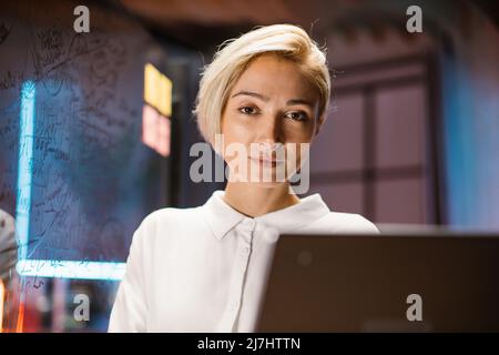 Gros plan sur une jolie femme d'affaires blanche blonde souriante en chemise blanche, posant à la caméra, dans un bureau sombre moderne. Arrière-plan intérieur de bureau flou, panneau de verre Banque D'Images