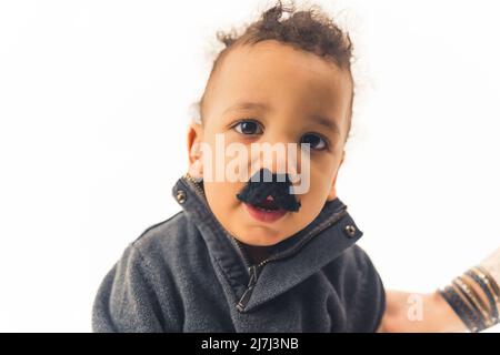 Portrait en studio d'un adorable bébé biracial avec des cheveux fclés foncés regardant l'appareil photo tout en portant une fausse moustache noire sur fond blanc. Photo de haute qualité Banque D'Images