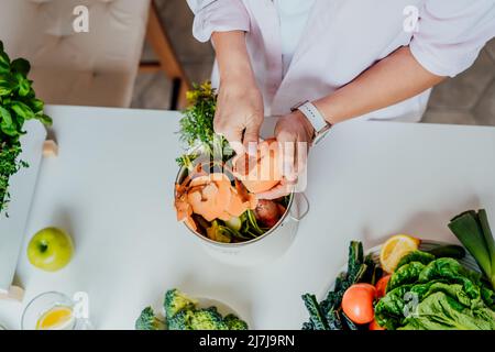 Composte les déchets de cuisine, recyclage à la maison. Vue de dessus la femme nettoie les patates douces et met les légumes coupés restes dans les ordures, bac de compost sur Banque D'Images