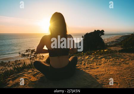 Femme pratiquant le yoga et médite sur la montagne avec vue sur l'océan au coucher du soleil. Une jeune femme assise à lotus pose à l'extérieur dans la nature Banque D'Images