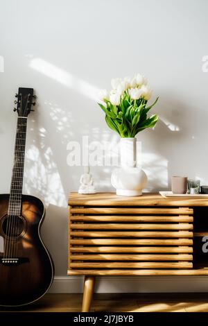 Décoration intérieure minimaliste scandinave - bouquet de fleurs de tulipe blanches dans un vase, bougies, statuette de Bouddha sur une armoire en bois. Plante de maison verte Banque D'Images